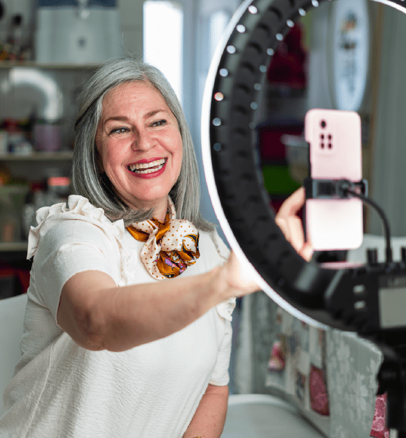 smiling woman with gray hair sits in a brightly lit room, posing for a selfie or video using her smartphone mounted on a ring light tripod. She wears a white blouse with ruffled sleeves and a colorful patterned scarf around her neck. The background features shelves with various items, suggesting a creative or personal workspace. The atmosphere is vibrant and cheerful.